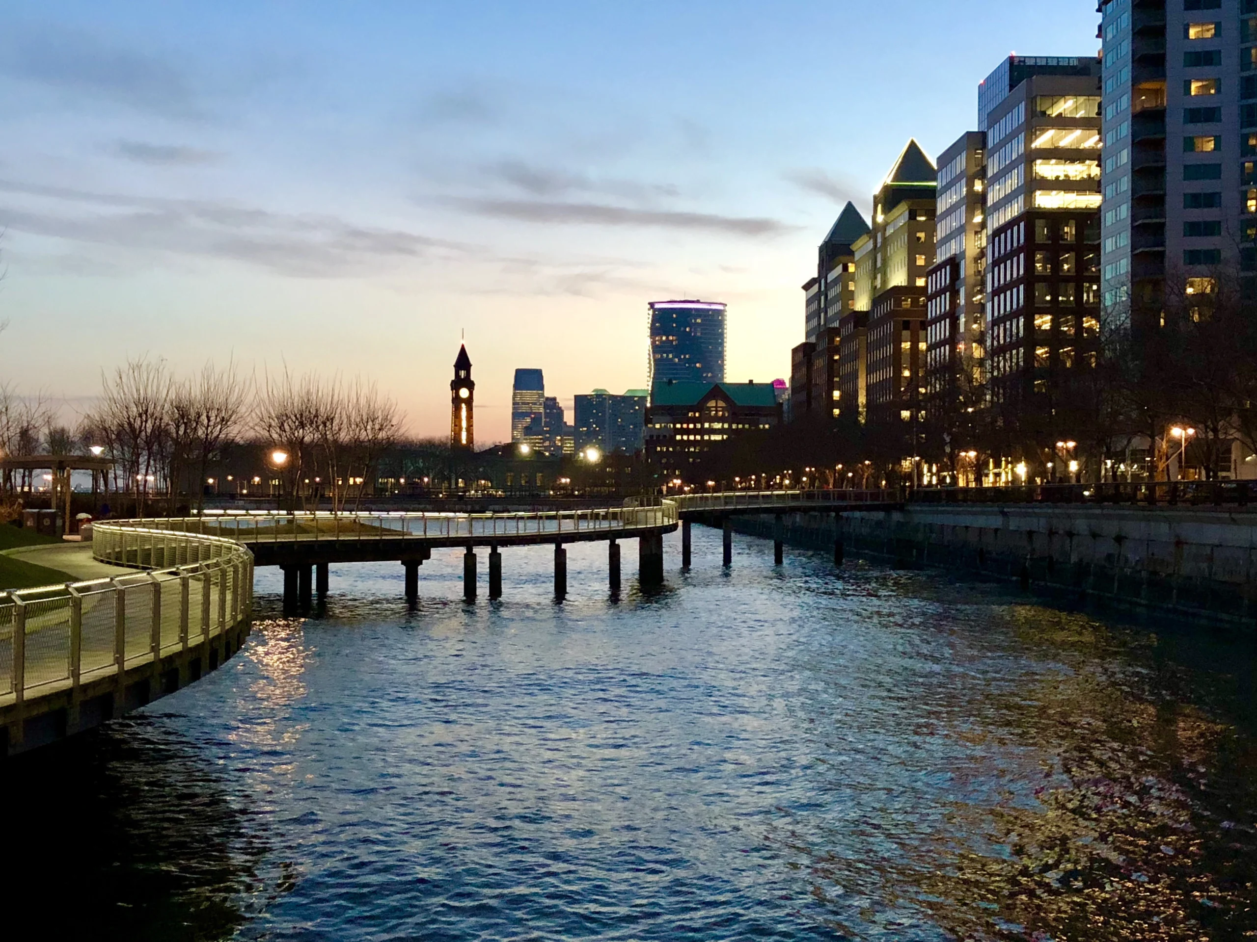 Waterfront boardwalk at dusk in Hoboken, New Jersey, with the Hoboken Terminal clock tower and the Jersey City skyline across the water